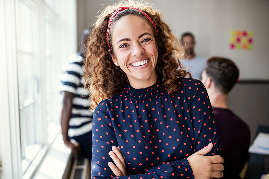 Employee Benefits - Portrait of a Young Smiling Employee Standing in a Meeting Room in the Office Next to a Window with Her Arms Folded