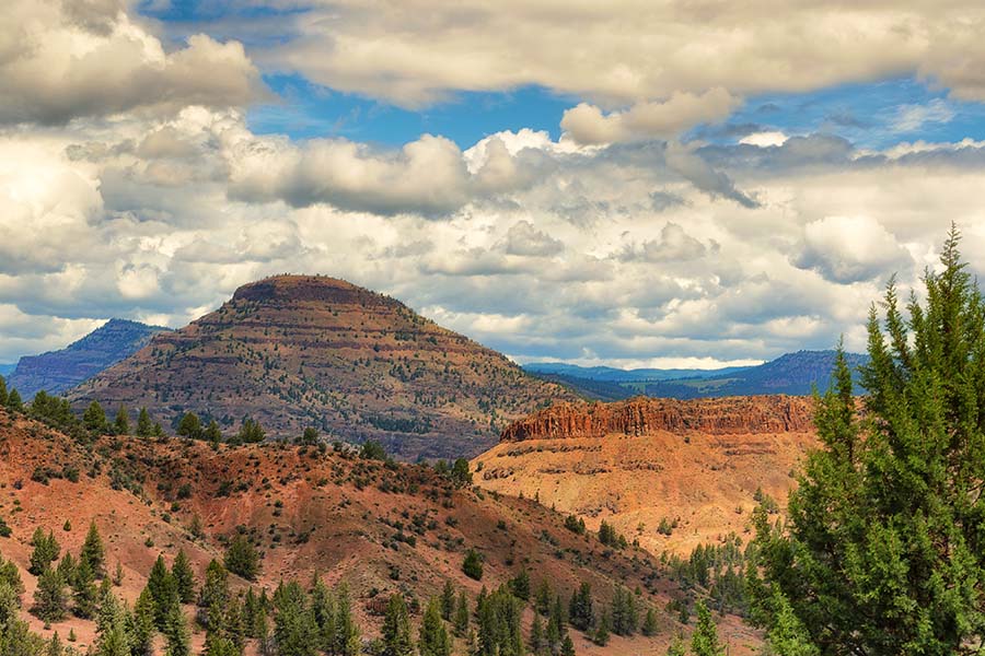Sheridan, OR - Scenic View of Red Sand Hills and Mountains with Green Trees Against a Cloudy Blue Sky in Sheridan Oregon